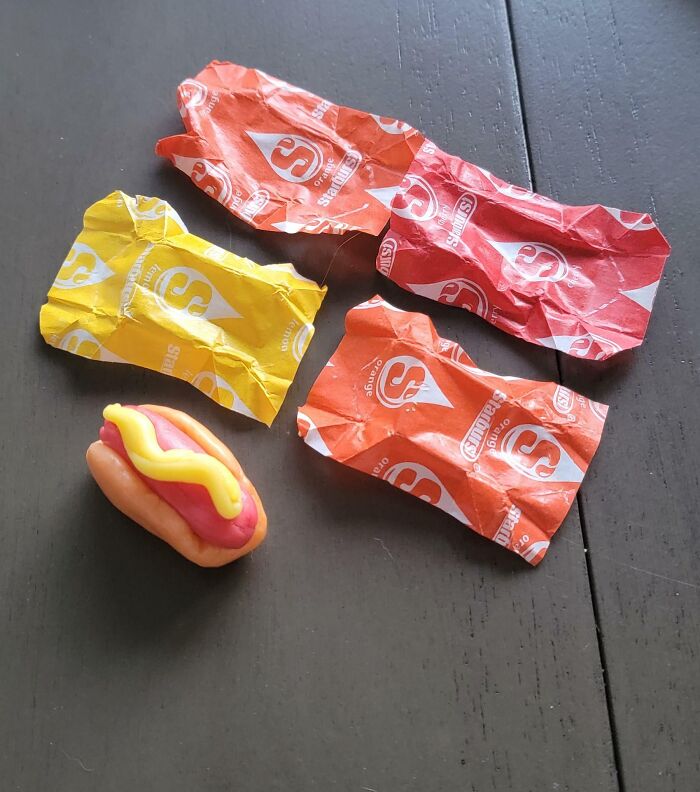 Small toy hot dog with mustard on a dark table next to four crumpled candy wrappers, showing fun sibling moments.