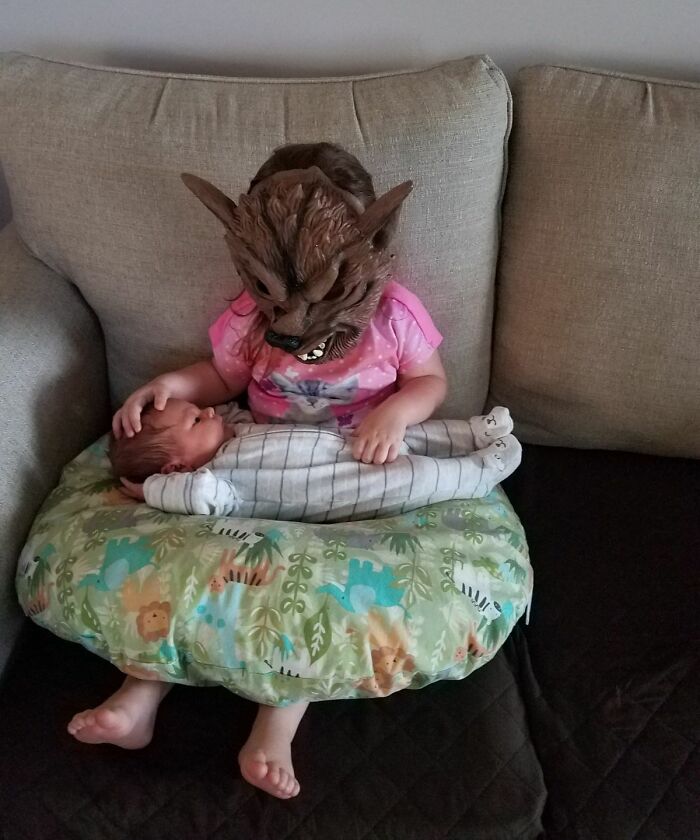 Toddler wearing a werewolf mask holding a newborn baby on a couch, capturing fun sibling moments and bond.