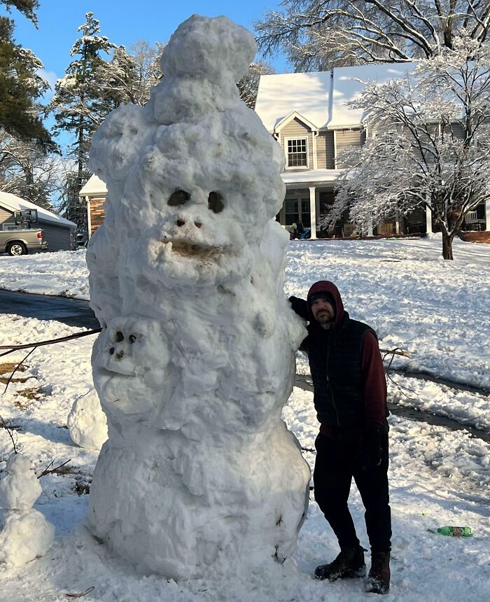 Man standing next to a huge snowman with multiple faces in a snowy yard, showing siblings having fun together outdoors.