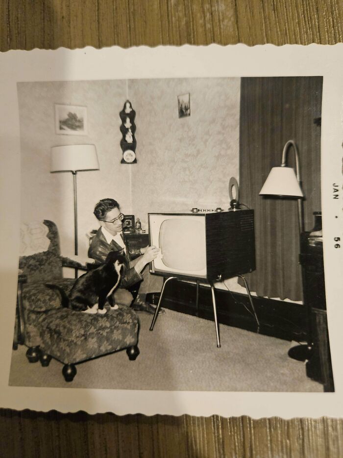Vintage photo of a man adjusting a 1950s TV with a black and white feline employee sitting on a chair nearby.
