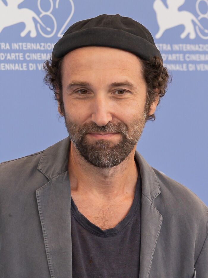 Thomas Bangalter wearing a black cap and gray jacket, attending an event with a blue background.