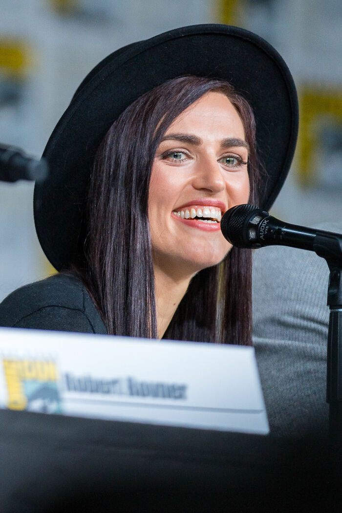 Katie McGrath smiling at a panel microphone, wearing a black hat and speaking at an event.