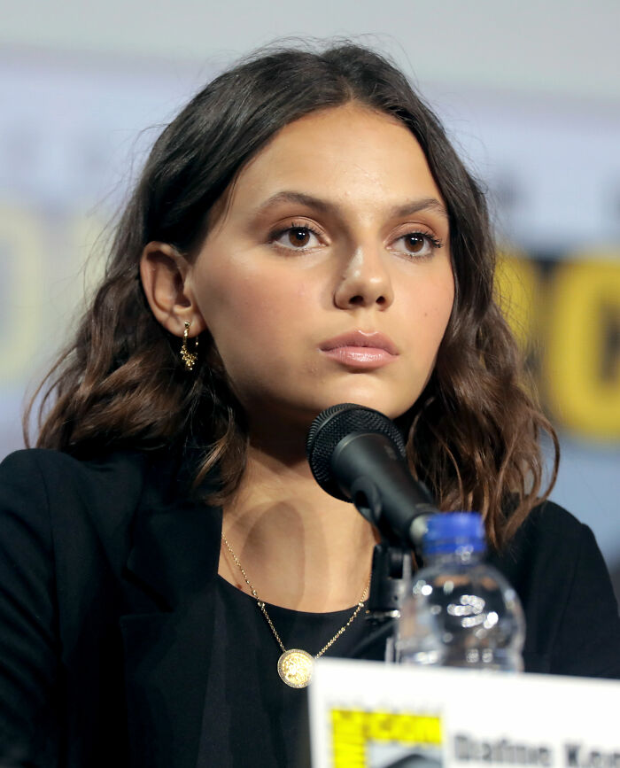 Dafne Keen speaking at a panel event, wearing a black outfit and gold jewelry with a focused expression.