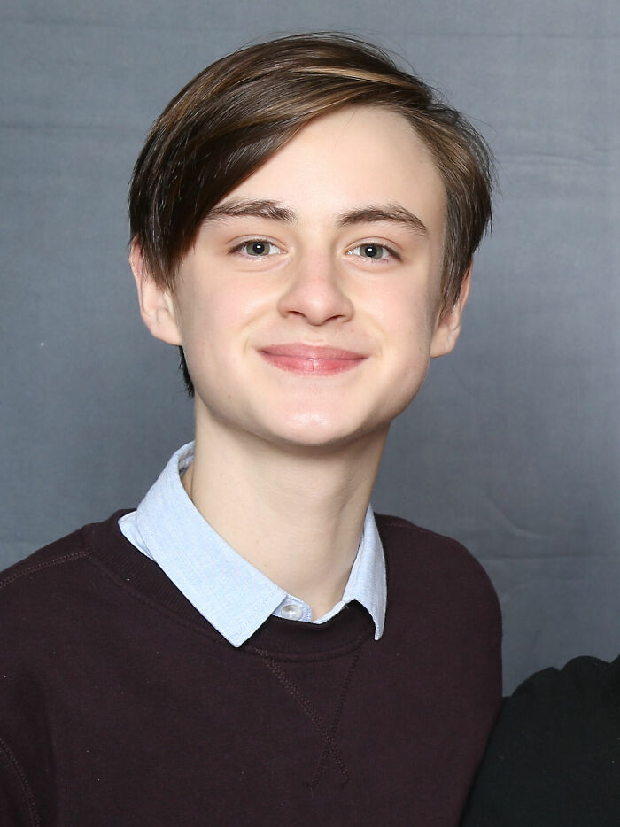 Jaeden Martell smiling, wearing a dark sweater over a collared shirt, posing against a plain gray background.