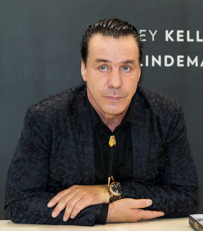 Till Lindemann seated at a table wearing a dark patterned jacket and bolo tie, with a focused expression.