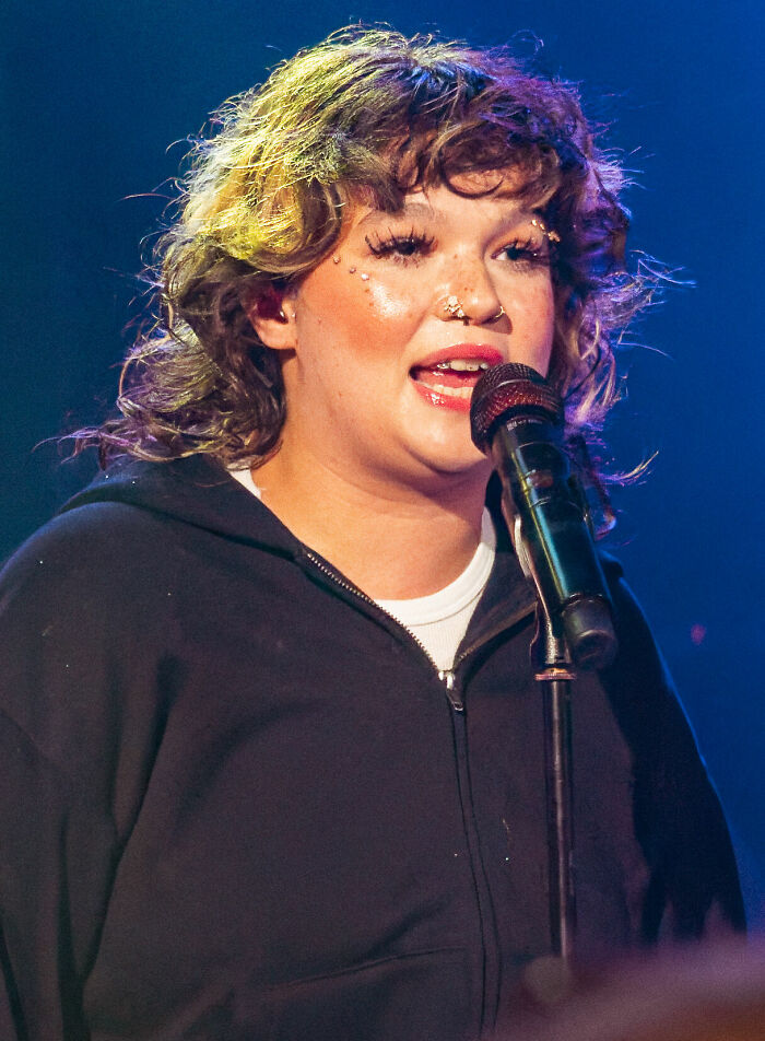 Lola Young performing on stage with curly hair and facial piercings, holding a microphone and wearing a black hoodie.