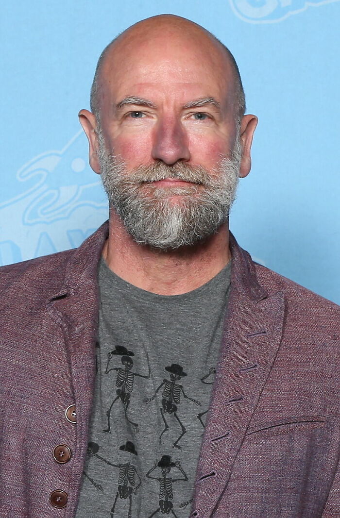 Graham McTavish with a trimmed beard wearing a gray skeleton print shirt and maroon blazer against a blue background