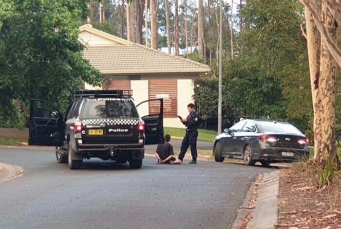Police officer detaining a person sitting on a suburban street beside a patrol SUV and parked sedan, next-level jerks