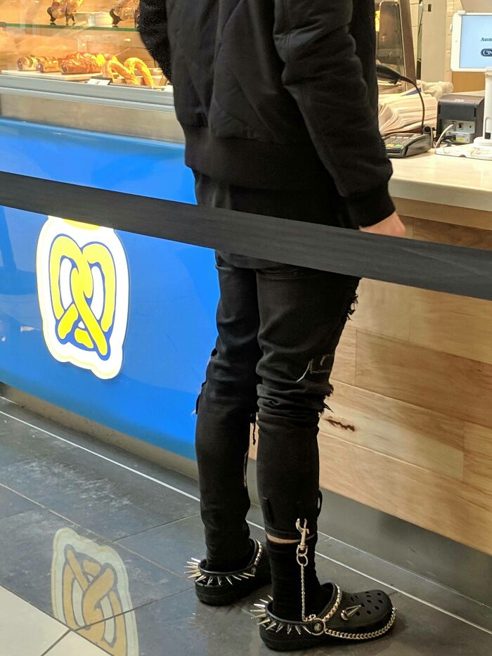Person wearing black ripped pants and spiked Crocs standing at a bakery counter, showcasing hilarious fashion fails. Person wearing black ripped pants and spiked Crocs standing at a bakery counter, showcasing hilarious fashion fails.