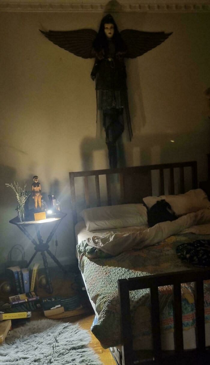 Figura oscura con alas en pared sobre cama en habitación tenue, evocando publicaciones espeluznantes que hacen cuestionar la realidad.