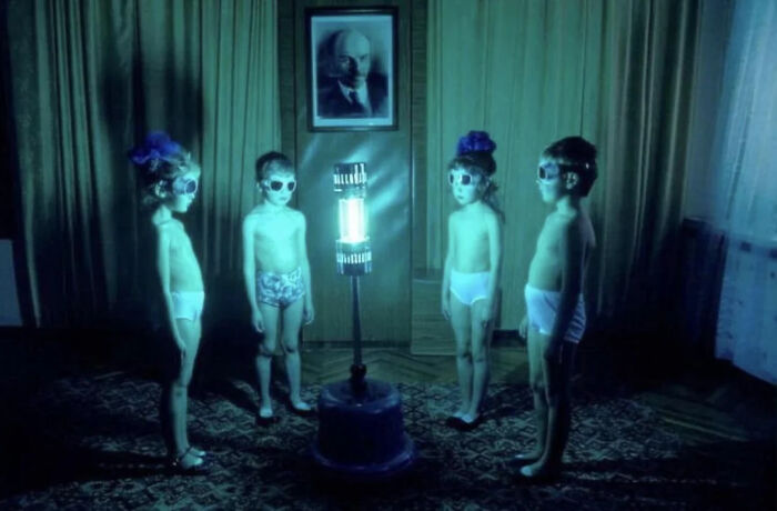 Four children wearing sunglasses and underwear stand around a glowing lamp in a dark room, eerie and unsettling scene.