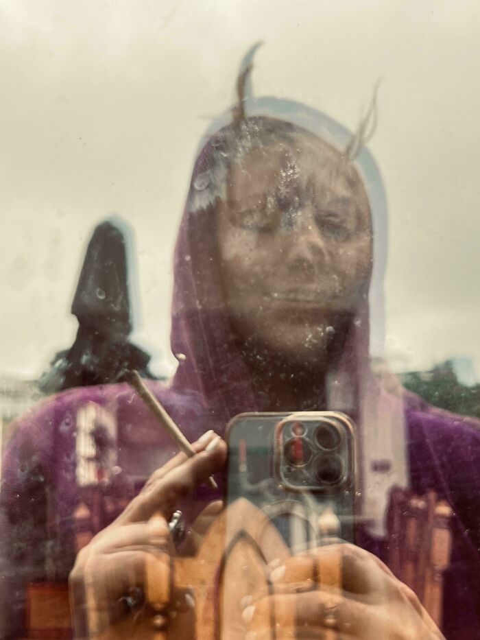 Reflection of a person in a blurred, dirty window holding a phone and cigarette, creating a scary post shared online effect.