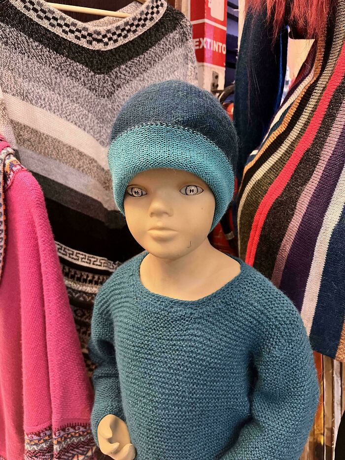 Mannequin with unsettling eyes wearing a blue knit hat and sweater among colorful scarves and sweaters in a store display.
