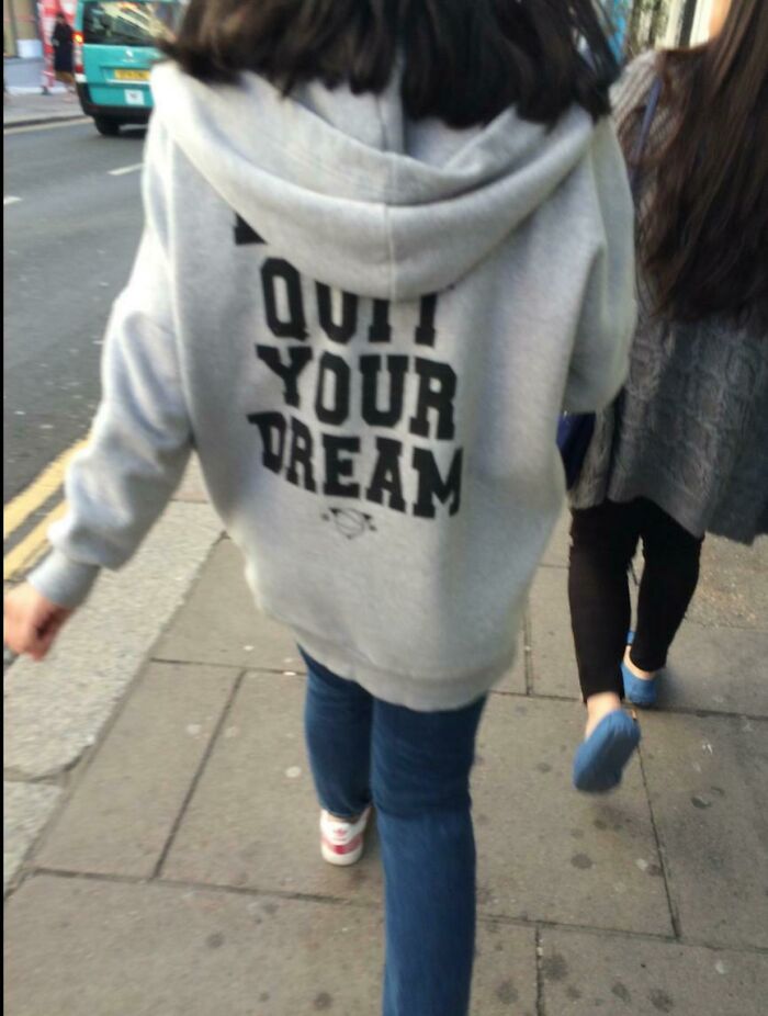 Person wearing a grey hoodie with a misprinted motivational message walking on sidewalk, showcasing hilarious fashion fails.