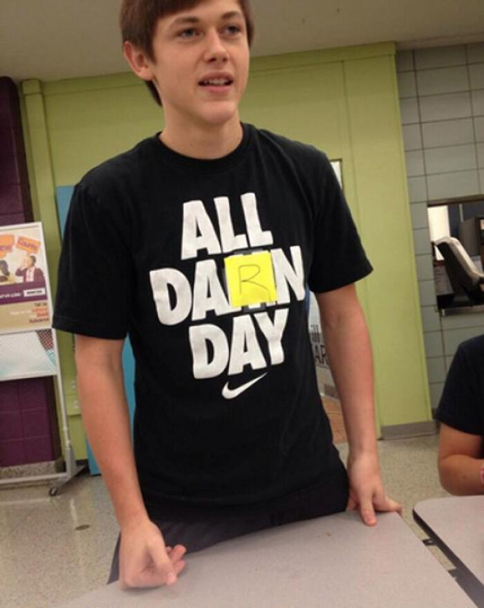 Teen wearing a black shirt with a taped letter creating a funny word mix-up, showcasing a hilarious fashion fail indoors.