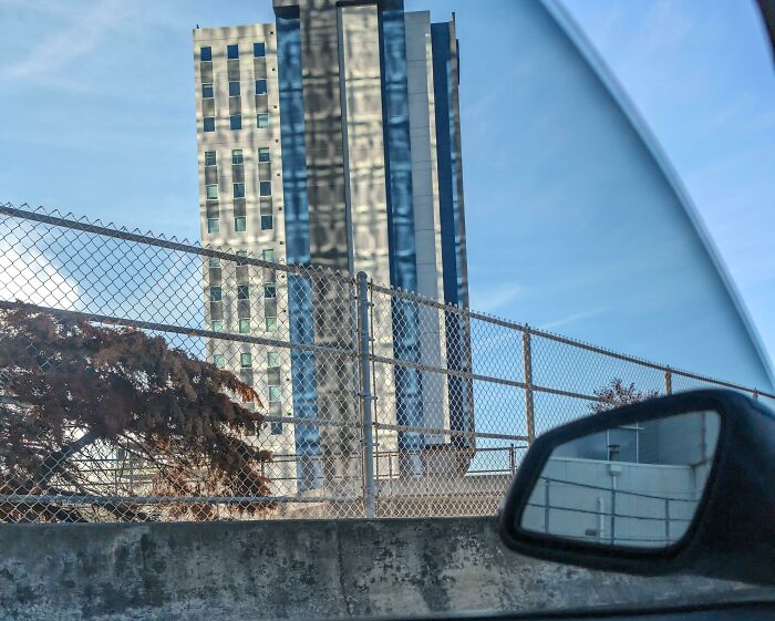 Car side mirror and chain-link fence framing a tall glass building with warped reflections, evoking glitches in the Matrix.
