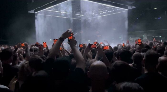 Concert crowd holding up glowing phones toward a large suspended light cube installation, glitches in the matrix vibe