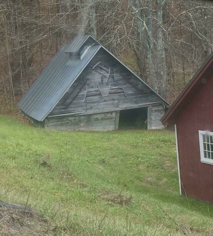 Leaning weathered wooden shed with strange markings beside red house on grassy slope, evoking glitches in the matrix