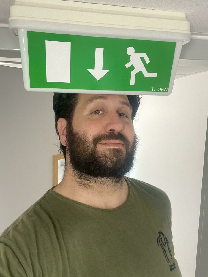 Man with beard in an olive shirt bumps head on a low ceiling sign, illustrating struggles only tall people face.