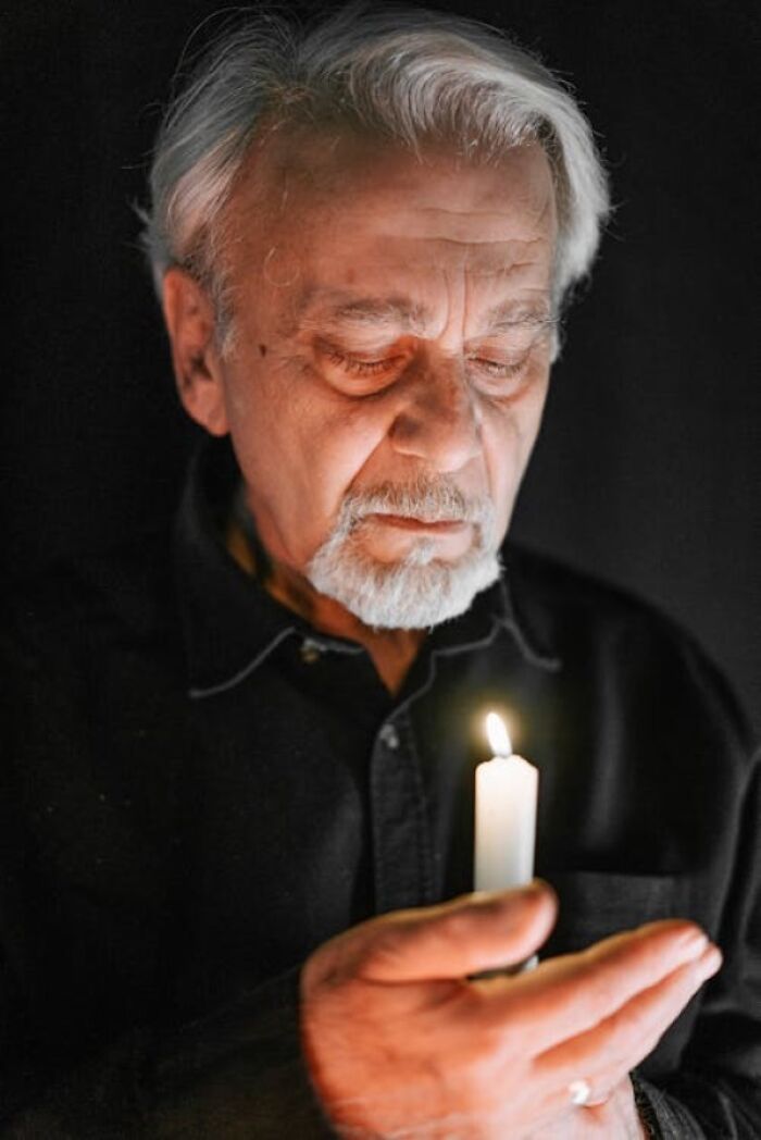 Elderly man holding a candle in darkness, reflecting quietly, evoking themes of family secrets and hidden stories.