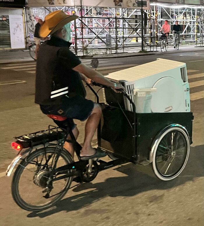 Man wearing a cowboy hat riding a cargo bike carrying a large appliance at night, showcasing impressive things countries implemented.