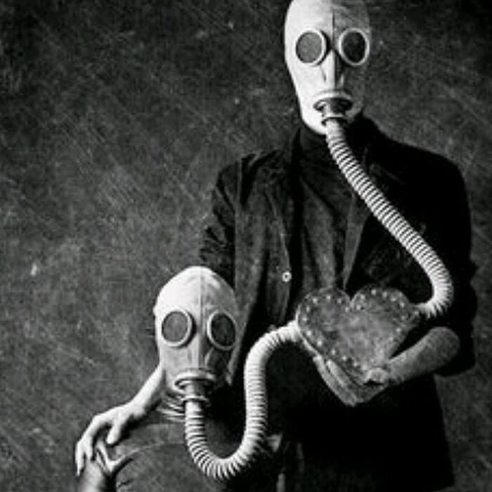 Two people wearing vintage gas masks connected by a hose, holding a heart-shaped object in an old black and white creepy photo.