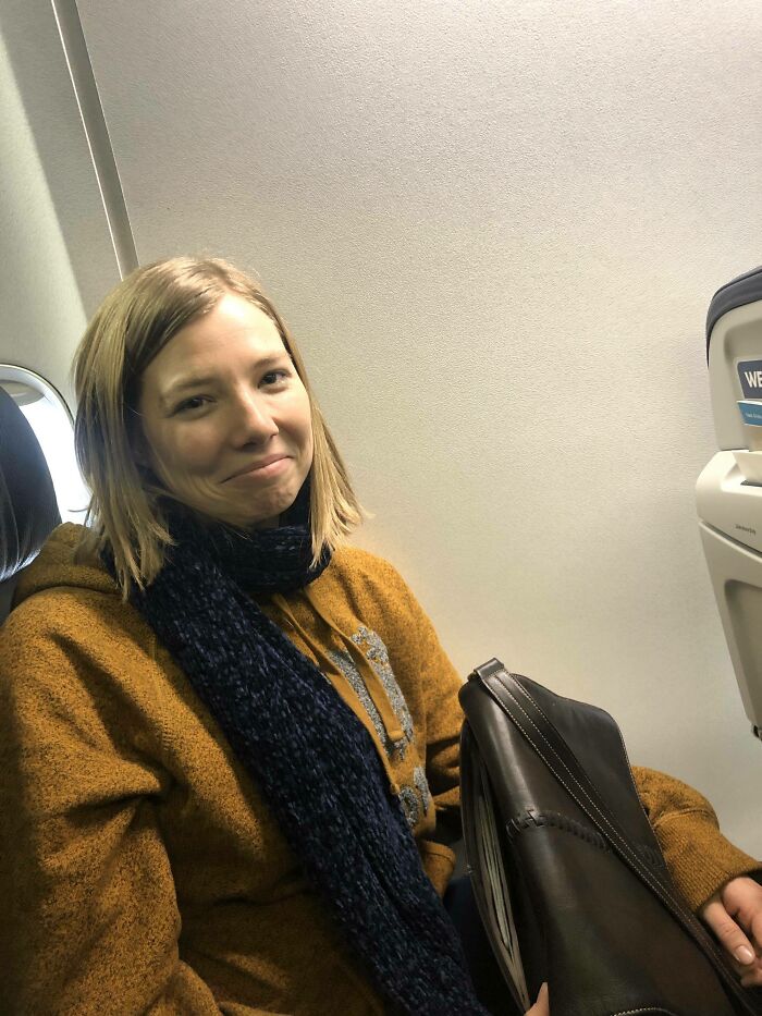 Woman on airplane with brown coat and navy scarf holding a bag, woke up expecting a normal day