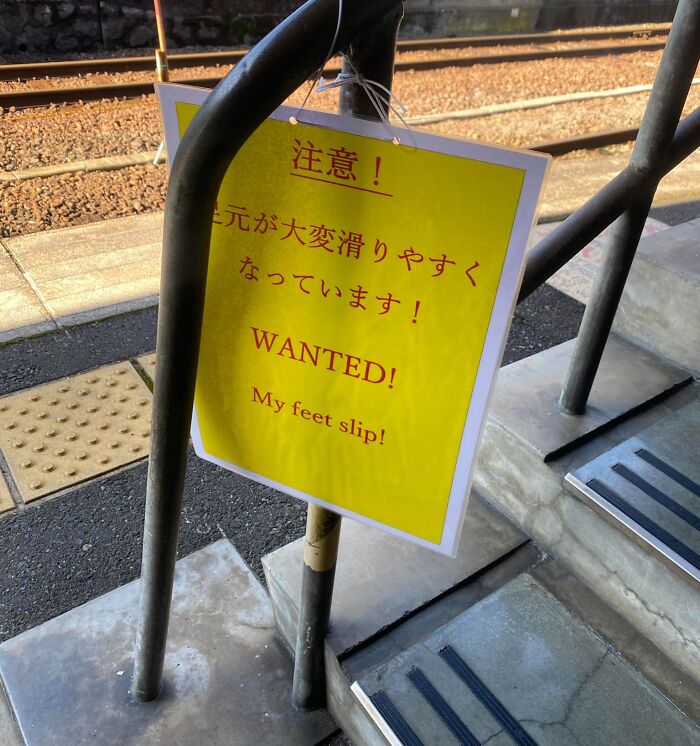 Yellow warning sign showing a funny Japan translation fail with the phrase "WANTED! My feet slip!" by train station stairs.