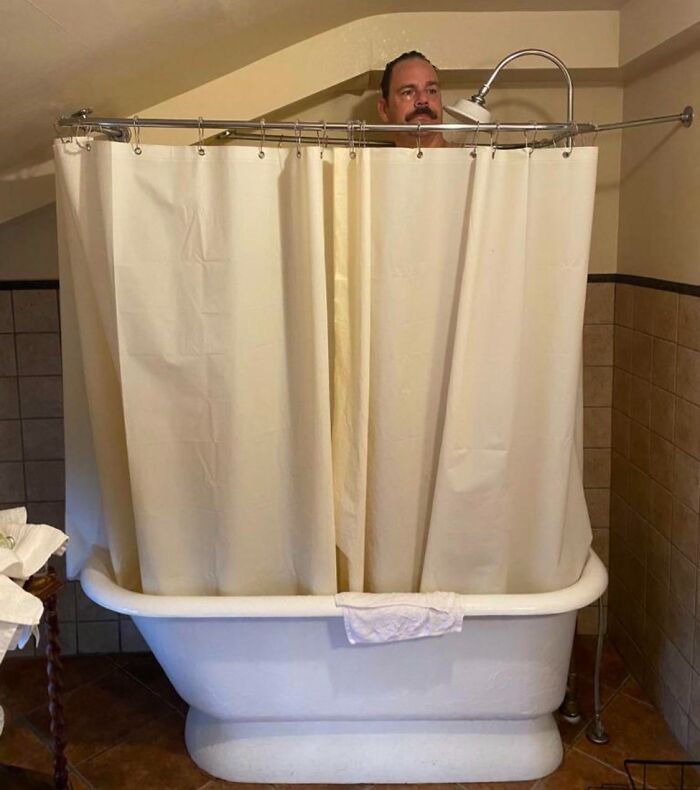 Man in a bathtub with shower curtain barely covering him, illustrating common struggles tall people face in bathrooms.