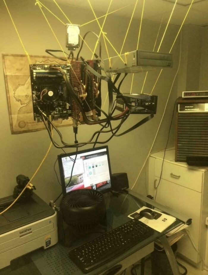 Confusing computer components suspended by yellow strings above a desk with a monitor and keyboard in an office setting.