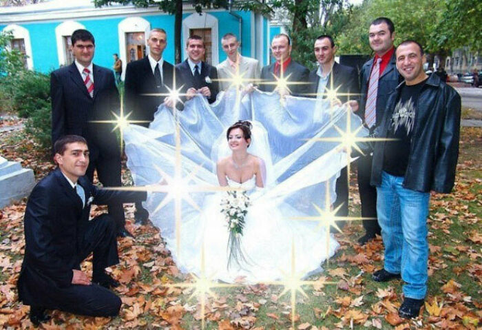 Bride sitting in a wedding dress surrounded by groomsmen holding fabric, creating an awkward wedding photo moment outdoors.