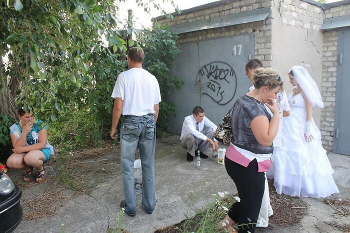 Awkward wedding photo showing a bride in white dress and guests in unusual poses outside a weathered garage door.