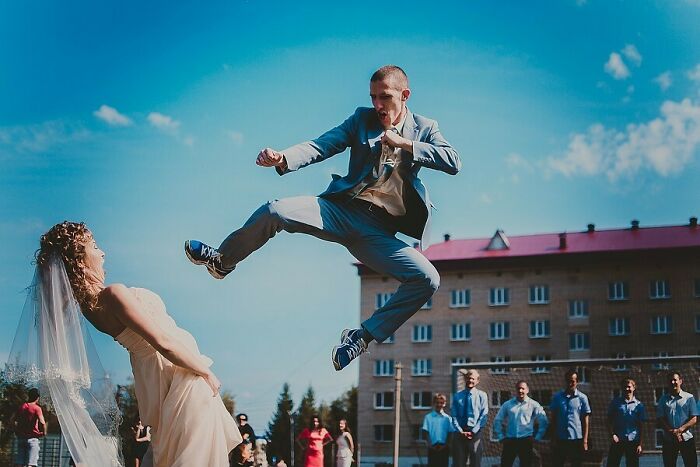Groom in mid-air performing a martial arts kick toward bride during an awkward wedding photo outside with guests watching.