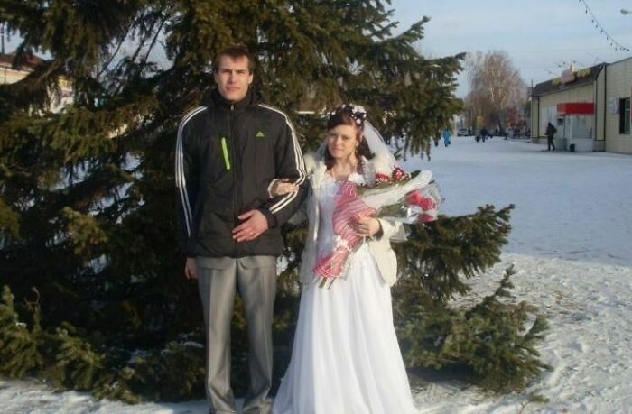 Bride in wedding dress holding bouquet next to groom in casual jacket, standing awkwardly outdoors in snowy setting.