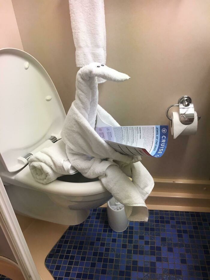 Towel animal shaped like a bird sitting on a toilet reading a brochure in a bathroom with blue tiles.
