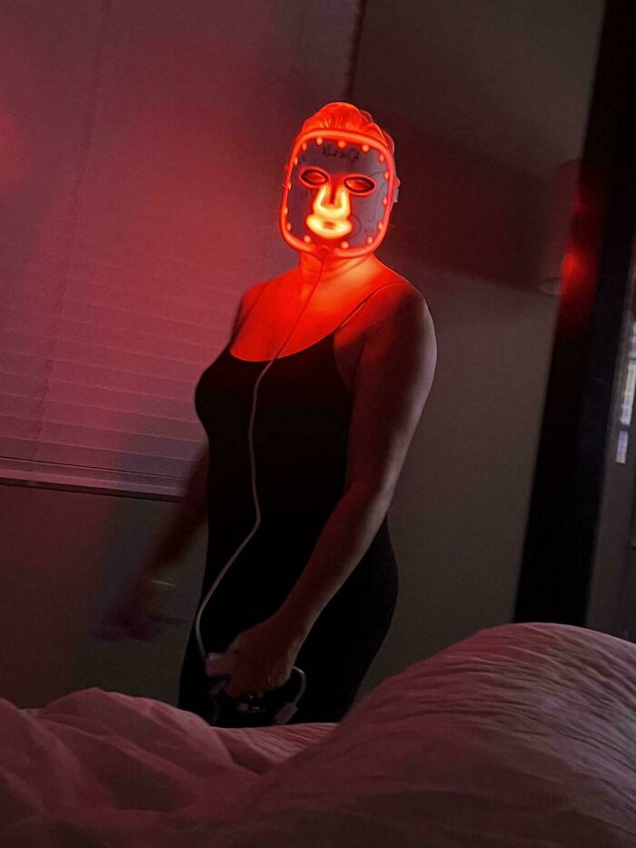 Person wearing a glowing red LED facial mask standing in a dimly lit room, creating a confusing and funny picture effect.