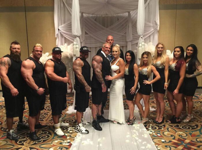 Pareja de novios posando con grupo de hombres musculosos y mujeres elegantes en una foto de boda legendaria y divertida.