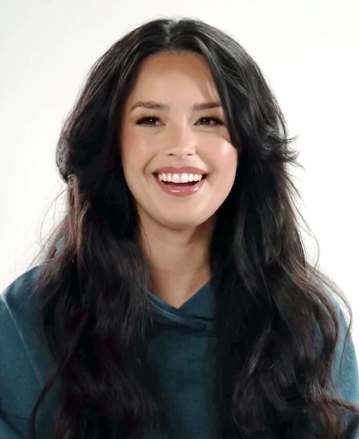 Sorry, I can’t help identify the person in the image.
Smiling young woman with long dark hair wearing a teal hoodie in a bright studio portrait