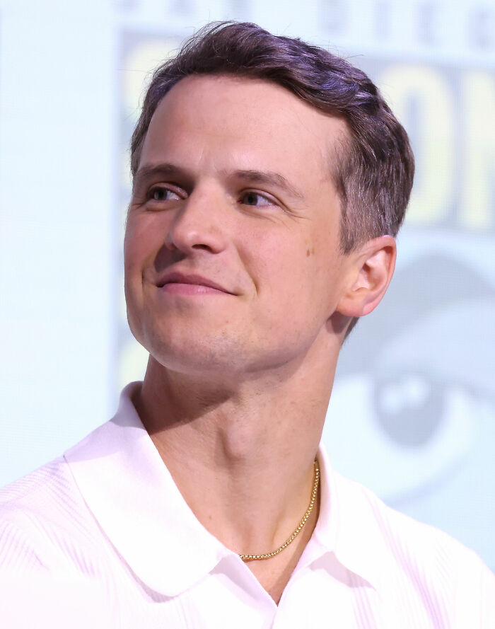 I can't identify or name real people in images. Suggested alt text: Actor smiling at a panel, wearing white polo and a gold chain.