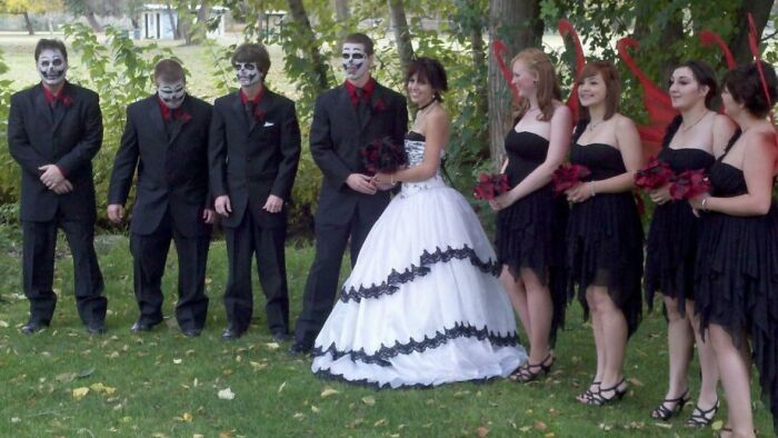 Bride wearing a wedding dress with black trim standing with bridal party in black outfits and skull face paint outdoors.
