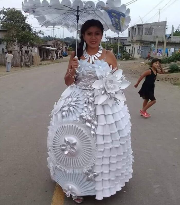 Woman wearing a wedding dress made from unconventional materials standing outdoors holding a large white umbrella.