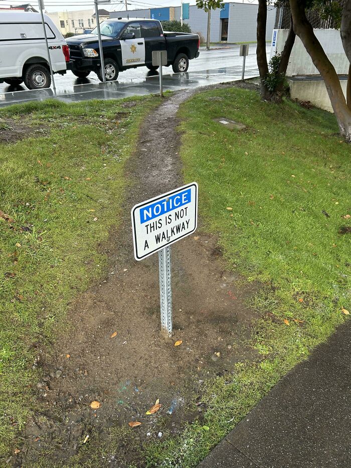 Desire path worn on grass with a sign stating this is not a walkway, showing how people walk in reality versus city planning.
