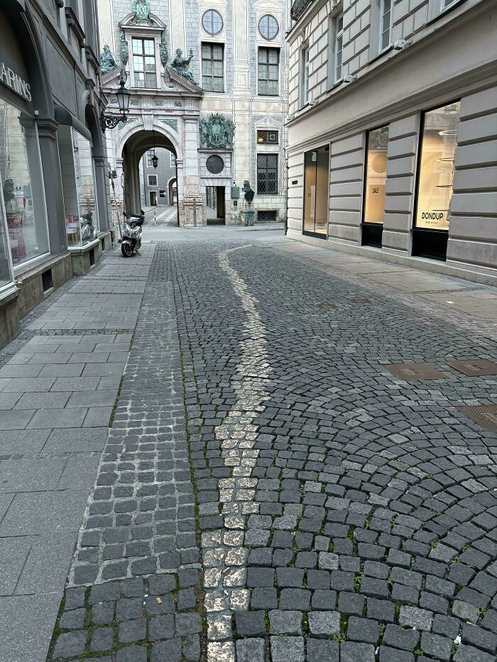Narrow desire path marked by worn stones curving through cobblestone street between city buildings and archway ahead.