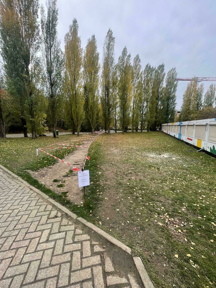 Desire path worn through grass in a city park, blocked off by tape and a sign, showing how people walk versus planned paths.
