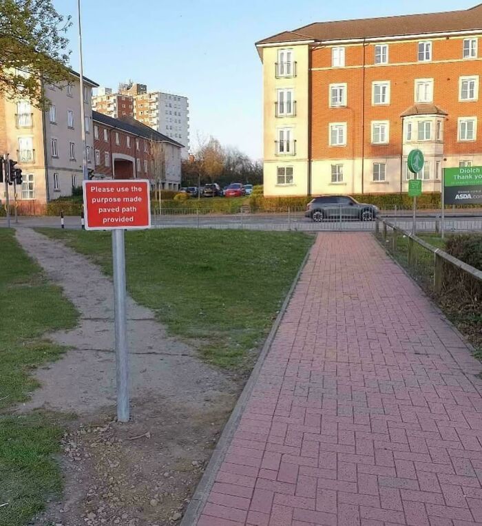A desire path formed on grass alongside a paved walkway with a sign instructing to use the paved path in a city setting.