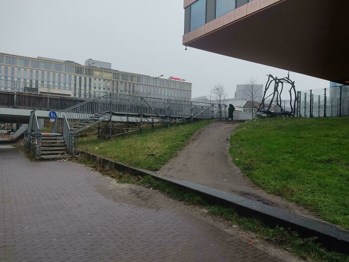 Desire path cutting diagonally across grass hill beside pedestrian stairs and modern buildings in an urban setting.