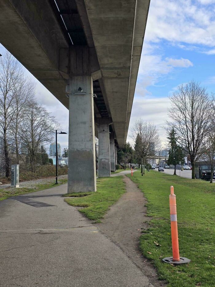 Concrete urban overpass with paved sidewalk and visible desire path worn into grass alongside it.