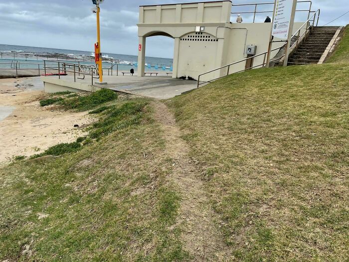 Desire path worn on grass beside concrete walkway near beach showing how people walk differently from city plans.