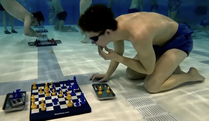 Young man playing chess underwater wearing goggles and swim trunks in a confusing and funny picture.
