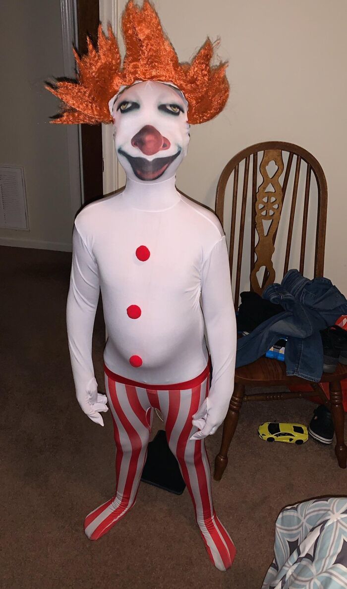 Child in a full-body clown costume with orange hair and striped pants, a hilarious fashion fails moment captured indoors.